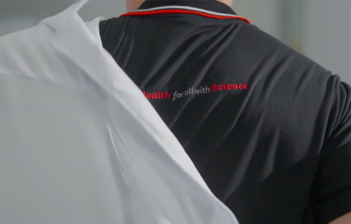 Back view of a health science professional in a black polo shirt with text, putting on a white lab coat.